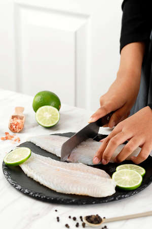 Female Hand Cutting Dori Fillet Pangasius Fish above Stone Board, Process Cooking in the Kitchenの写真素材