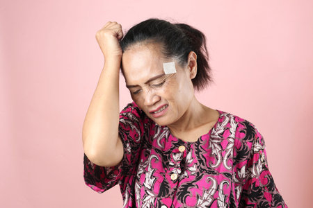Indonesian Asian Woman Wearing Daster Home Dress with Hot Patch, Holding Her Head Having Headacheの写真素材