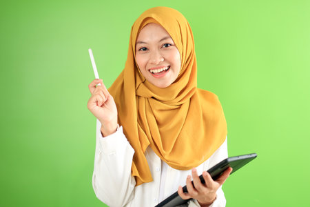 Asian Hijab Girl Holding Tab and Pen, Smiling at the Camera, Isolated on Green Backgroundの写真素材