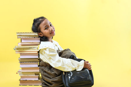 Asian Kids Girl Sitting and Leaning on Stacked Books, Isolated on Yellow Backgroundの写真素材