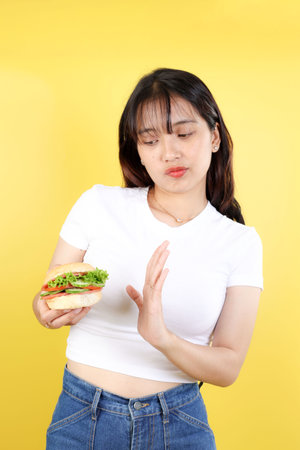 Young Sad Asian Woman Wear White Clothes Hold Eat Burger Show Hand Stop Gesture Say No, Isolated on Yellow Background. Proper Nutrition Healthy Lifestyle Fast Food Unhealthy Choice Conceptの写真素材
