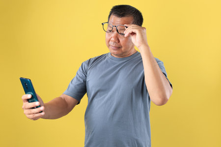 Middle Aged Indonesian Man Squinting at Smart Phone, Senior Asian Man with Presbyopia. Isolated on Yellow Studio Backgroundの写真素材