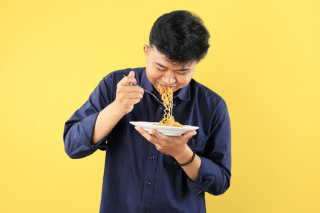 Young Asian Man Looking Down Enjoy Instant Noodles with Happy Face. Isolated on Yellow Background.の写真素材