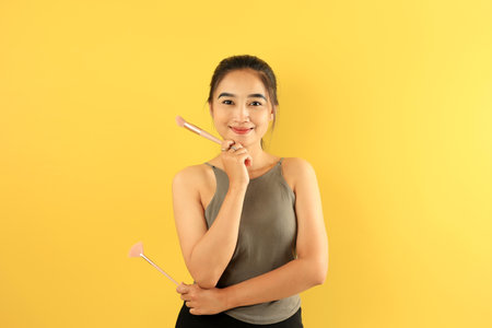 Asian Young Woman Holding Make Up Brush Smiling to the Camera, Isolated on Yellow Backgroundの写真素材