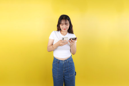 Portrait of a Happy Young Asian Girl Holding Credit Card while Holding Mobile Phone Isolated Over Yellow Backgroundの写真素材
