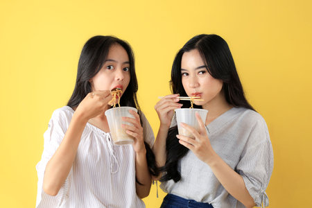 Two Young Asian Girl Holding A Cup of Noodle, Isolated on Yellow Background. Copy Space for Text