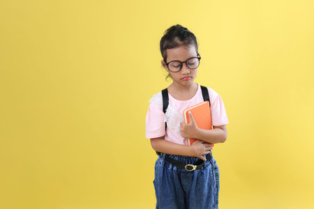 Asian Little Girl Kid Pouting with Backpack and Book. Isolated on Yellow Backgroundの写真素材