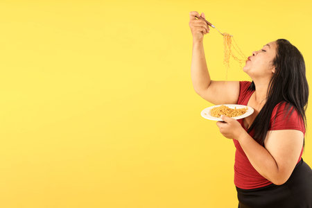 Indonesian Fat Woman Eating Indomie Goreng Instant Noodle, Isolated on Yellow Background, Copy Spaceの写真素材