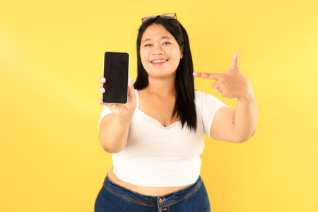 Asian Fat Big Size Woman Pointing at Mobile Phone, Isolated on Yellow Backgroundの写真素材