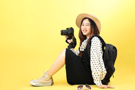 Young Asian Girl Backpacking Concept with Camera and Hat Straw, Isolated on Yellow Backgroundの写真素材