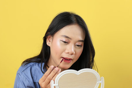 Pretty Asian Young Woman, Girl Make Up Face by Applying Red Lipstick, Lips Balm on her Lips using Lip Applicator. Looking at The Mirror, Isolated on Yellow Background. Beauty Cosmetics Conceptの写真素材
