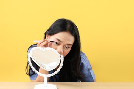 Young Asian Woman Applying Make-up to Her Face, using Black Eyeliner for Eyes and Looking at the Mini Standing Mirror. Isolated on Yellow Backgroundの写真素材
