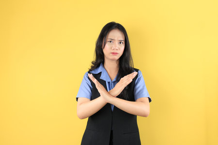 Young Indonesian Woman with Black Vest Blue Shirt, Making Rejection No Gesture with Crossed Her Arm Hand Forward, Isolated on Yellow Studio Backgroundの写真素材