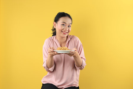 Asian Young Woman in Peach Shirt Holds a Plate of Mie Goreng Noodles with Happy Excited Face. She Sitting Againt Yellow Studio Backgroundの写真素材