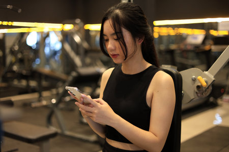 Young Female Sitting on Workout Bench in the Gym While Holding Mobile Phone, Watching Videoの写真素材