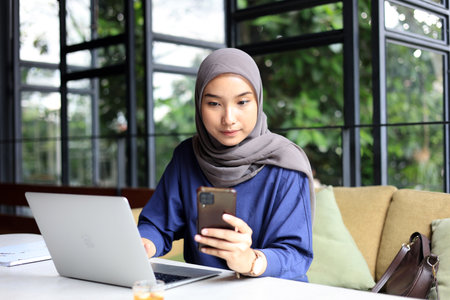 Young Asian Muslim Business Woman Wearing Hijab using Smartphone with Laptop, Sitting at a Cafe...