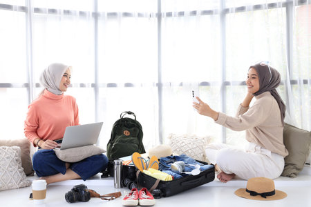 Two Asian Young Female Hijab Muslim Friends Preparing for Summer Trip, Book Tickets using Laptop and Taking Selfie Picture for Outfit of the Dayの写真素材