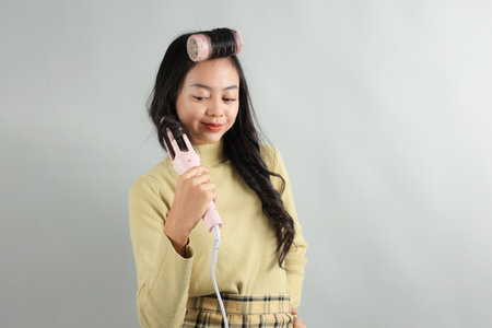 Beautiful Young Asian Teenager using Curling Hair Iron on Grey Background. Hair Styling Concept. Copy Space for Textの写真素材