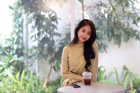 Attractive Young Asian Woman Smiling Behind a cup of Ice Coffee Americano on a White Table. A portrait of a Beautiful Indonesian Female Teenager Ssmiling Brightly at the Camera.の写真素材