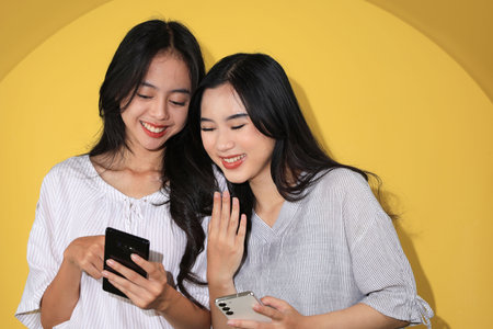 A Cheerful Asian Young Woman Showing Some Funny Video Meme to Her Female Friend via Modern Smartphone. Woman Bondings Interaction, Isolated on Yellow Backgroundの写真素材