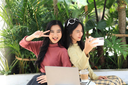Asian Woman Friends Using Smartphone Selfie Together while Sitting at Outdoor Coffee Shop. Drinking Coffee in Urban City Street. Beautiful Female Enjoy Outdoor Lifestyle in the Cityの写真素材