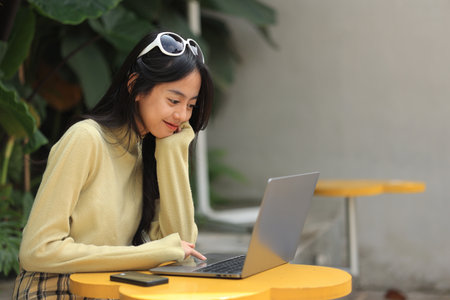 Happy Asian Young Girl Smiling and Working on Laptop Screen on Outdoor Coffee Shop with Green Plant and Yellow Small Tableの写真素材