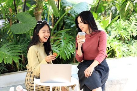 Two Young Asian Woman Chatting and Joyful with Food  and Coffee in a Coffee Shop Restaurant. Working with Laptop Togetherの写真素材