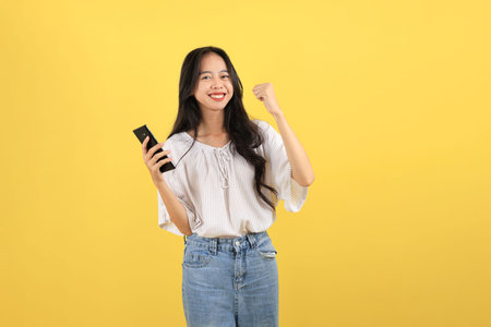 Young Indonesian Teenager with Long Hair Smiling with One Hand Raised While using a Black Smartphone, Isolated on Yellow Backgroundの写真素材