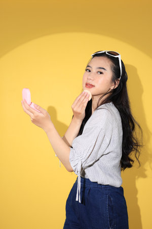 Stylish Young Asian-Caucasian Female Touch Up her Make Up using Pink Case Make Up Cushion, Looking at Camera. Isolated on Yellow Backgroundの写真素材