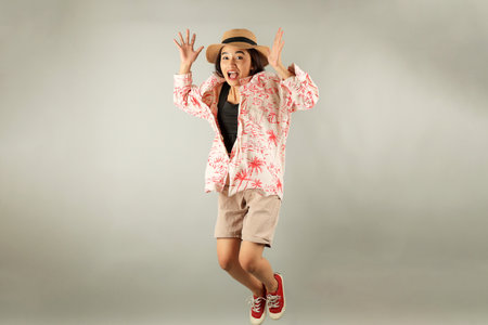 Young Asian Female Tourist Smiles at Camera and Jumps, Excited About Summer Vacation. Isolated on Grey Backgroundの写真素材