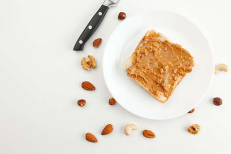 Bread with peanut butter on a plate. Knife and various types of nuts. Cashew, almond, walnut, hazelnut isolated on white background.の写真素材