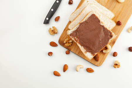 Square bread for toast with chocolate paste on a wooden board. Nuts, a knife and a wooden cutting board with a sandwich and slices of bread on a white table.の写真素材