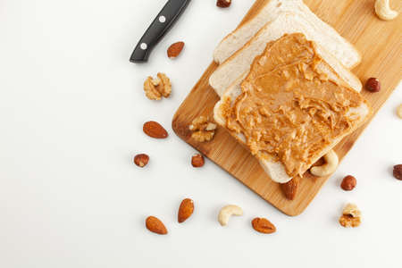 Square bread for toast with peanut butter on a wooden board. Nuts, a knife and a wooden cutting board with a sandwich and slices of bread on a white table.の写真素材