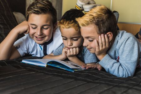 Focused little boys lying on bed and reading interesting story in book while spending free time...