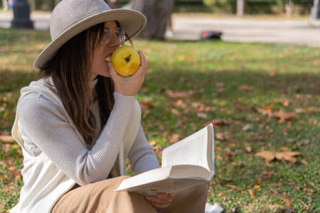 Brown woman in white hat, eats an apple while reading a book sitting in a park
