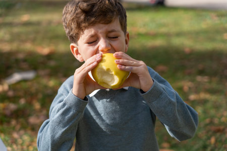 Boy in gray sweatshirt biting fresh apple with closed eyes while spending sunny weekend day in autumn parkの写真素材