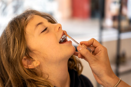 Cheerful girl closing eyes and smiling while crop makeup artist smearing lipstick on lips in beauty...
