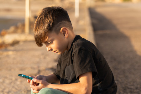 Boy in casual clothes sitting on border and watching video on cellphone on weekend day on beachの写真素材