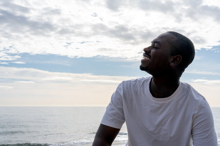 Positive African American male in white casual clothes smiling and looking away while sitting on beach near waving sea against cloudy sunset skyの写真素材