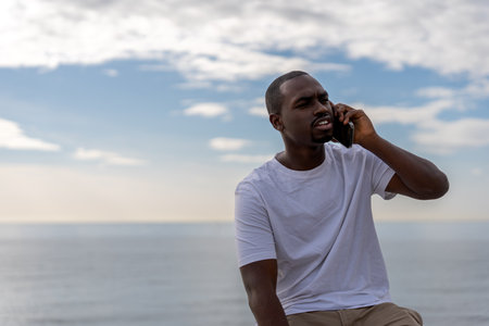 Serious African American male in casual clothes talking on cellphone while sitting against sea under skyの写真素材
