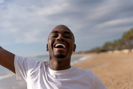 Cheerful African American male in casual clothes laughing and enjoying summer vacation on beach against cloudy skyの写真素材