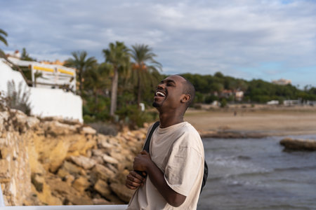 Happy African American male tourist in white t shirt with backpack laughing at funny joke while standing on terrace near waving seaの写真素材