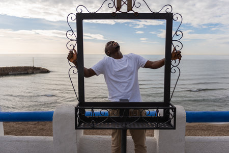 Laughing African American male in casual clothes standing at metal frame while enjoying vacation...