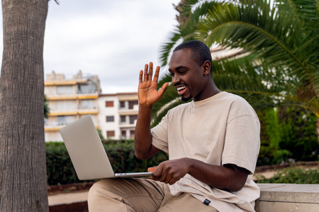 Cheerful African American male in casual clothes waving hand during video call via laptop at park