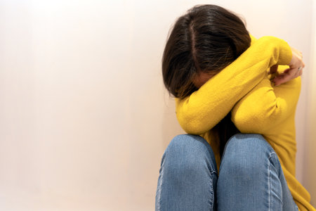 Anonymous woman in yellow sweater and jeans crying while sitting near beige wall and covering face...