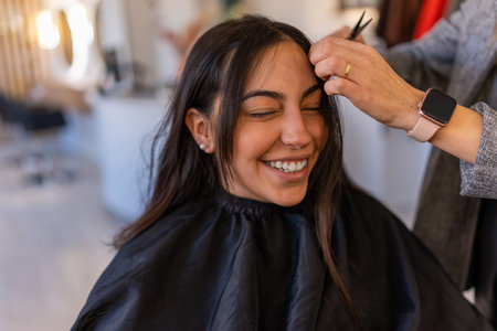Positive young female client smiling and sitting with closed eyes while getting hairstyle in...
