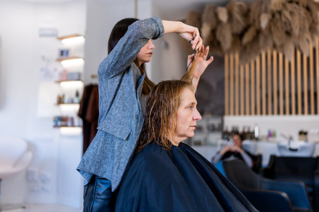 Side view of professional hairstylist cutting hair with scissors while preparing senior client for...