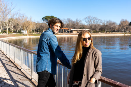 Young man leaning on railing of embankment while standing with mother wearing coat with hand in...