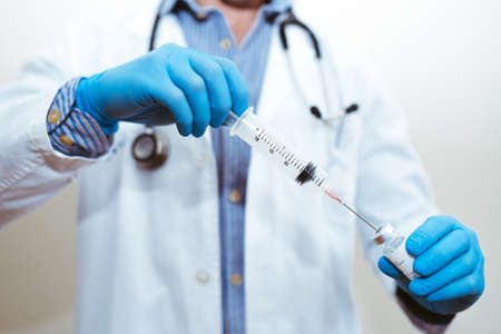 Doctor with gloves and uniform filling a syringe with liquid from a glass jar with a white backgroundの写真素材