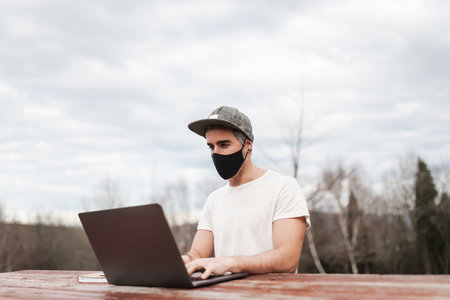 Man in a mask sitting in a table while working with a laptop in the middle of a fieldの写真素材
